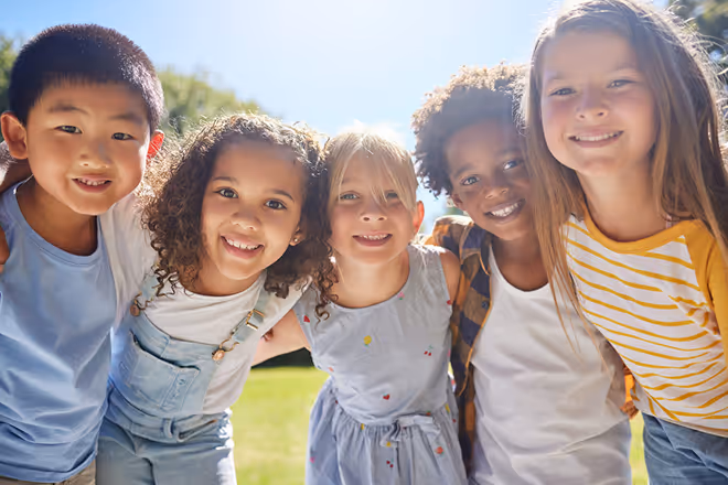 Group of five diverse children smiling and standing close together outdoors on a sunny day.