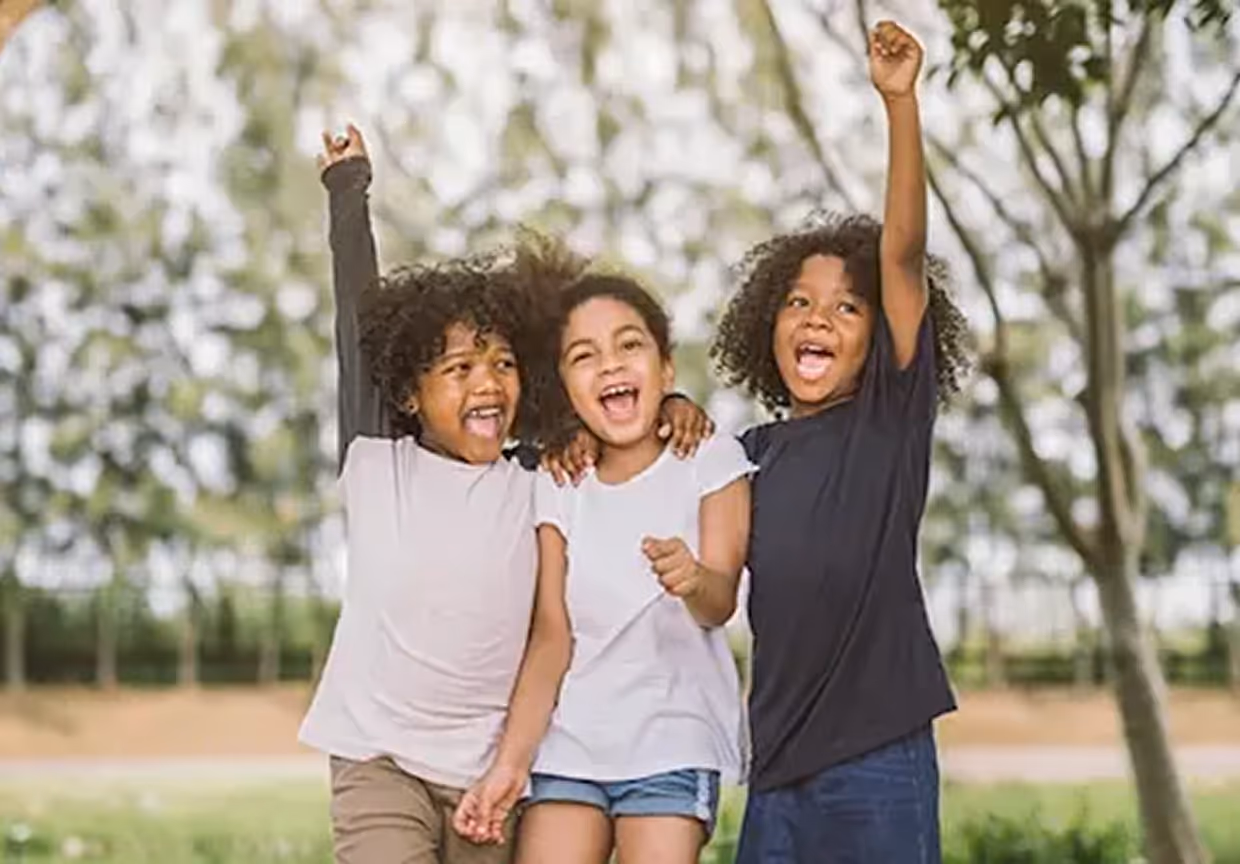 Three joyful children with curly hair smiling and raising their arms outdoors with trees in the background.