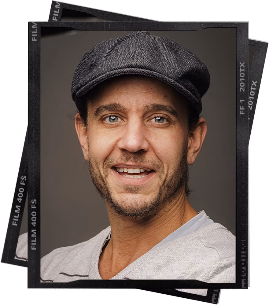 Smiling man with light stubble wearing a gray flat cap and a beige sweater against a gray background.