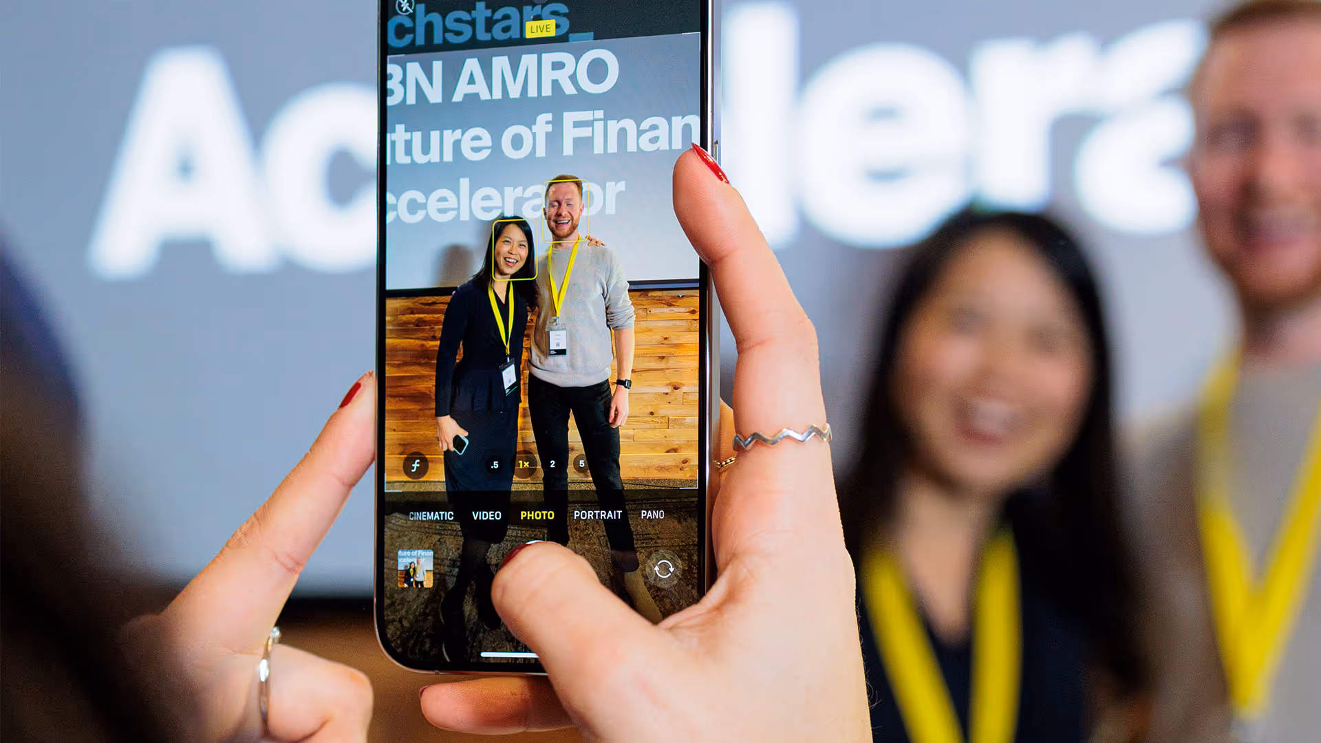 Two smiling people wearing yellow lanyards pose for a photo captured on a smartphone at a financial accelerator event.