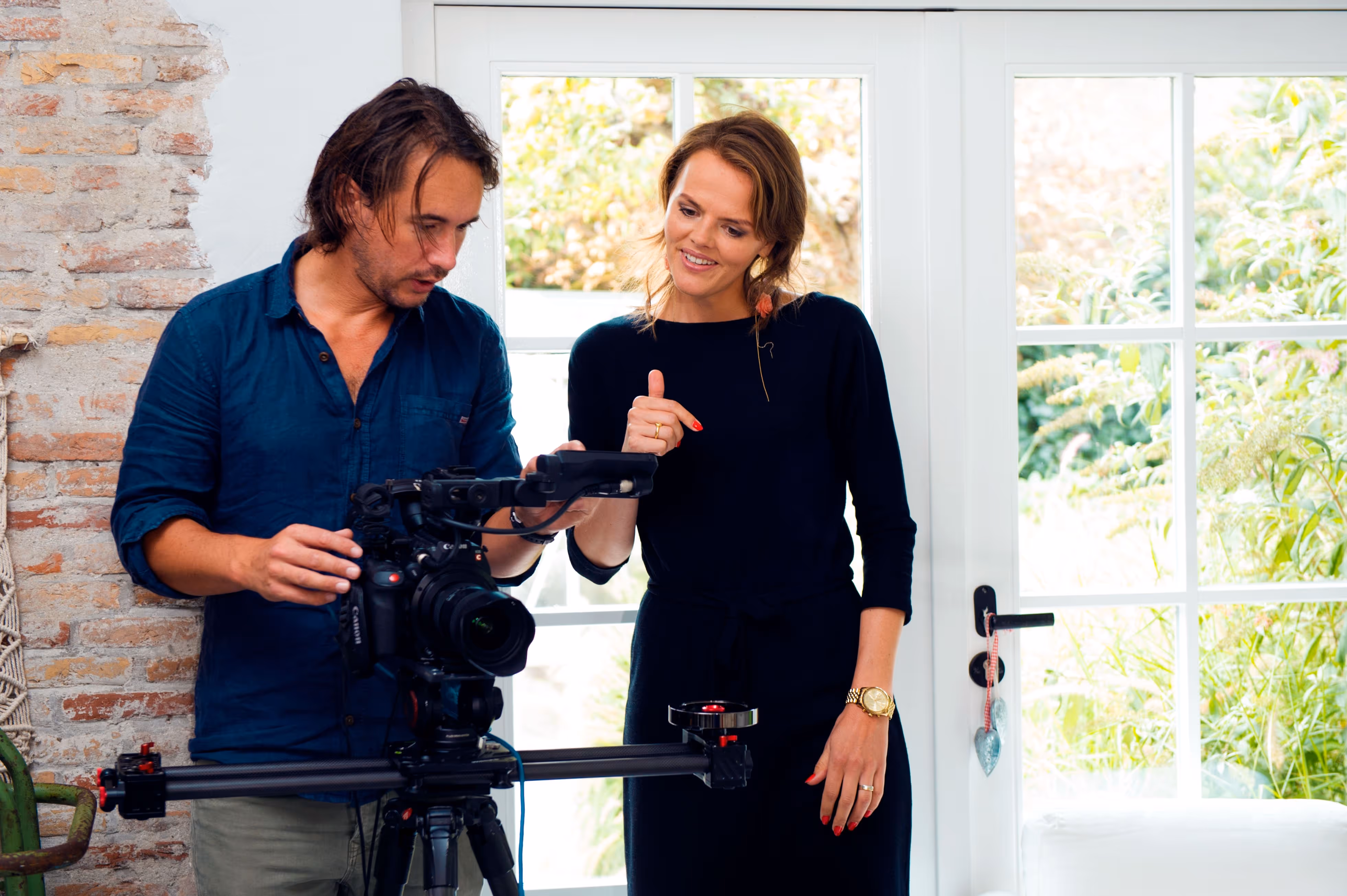 Man and woman operating a professional video camera on a slider in a bright room with large windows.