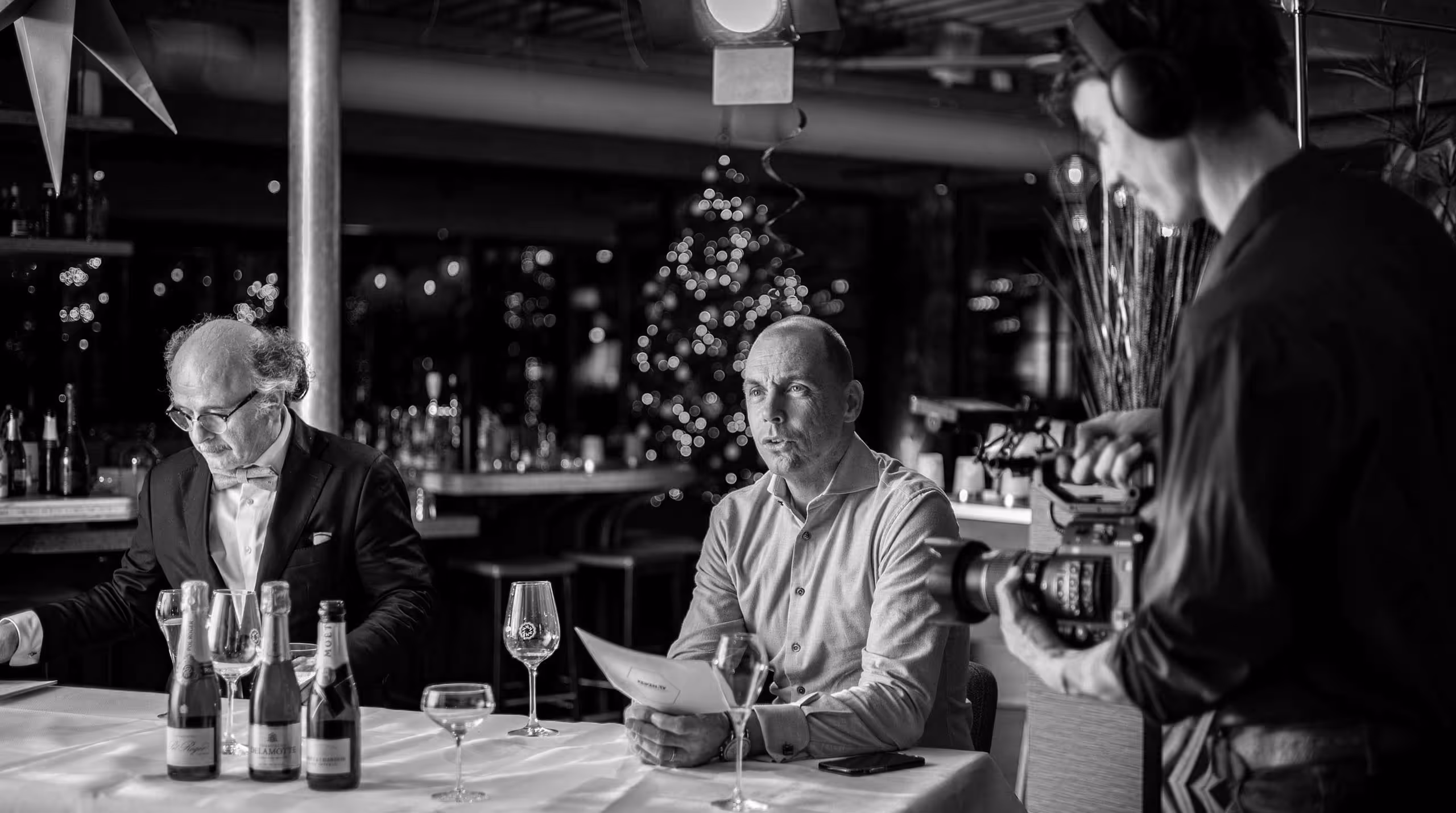 Black and white photo of two men seated at a table with bottles and glasses, while a cameraman films them in a decorated indoor setting.