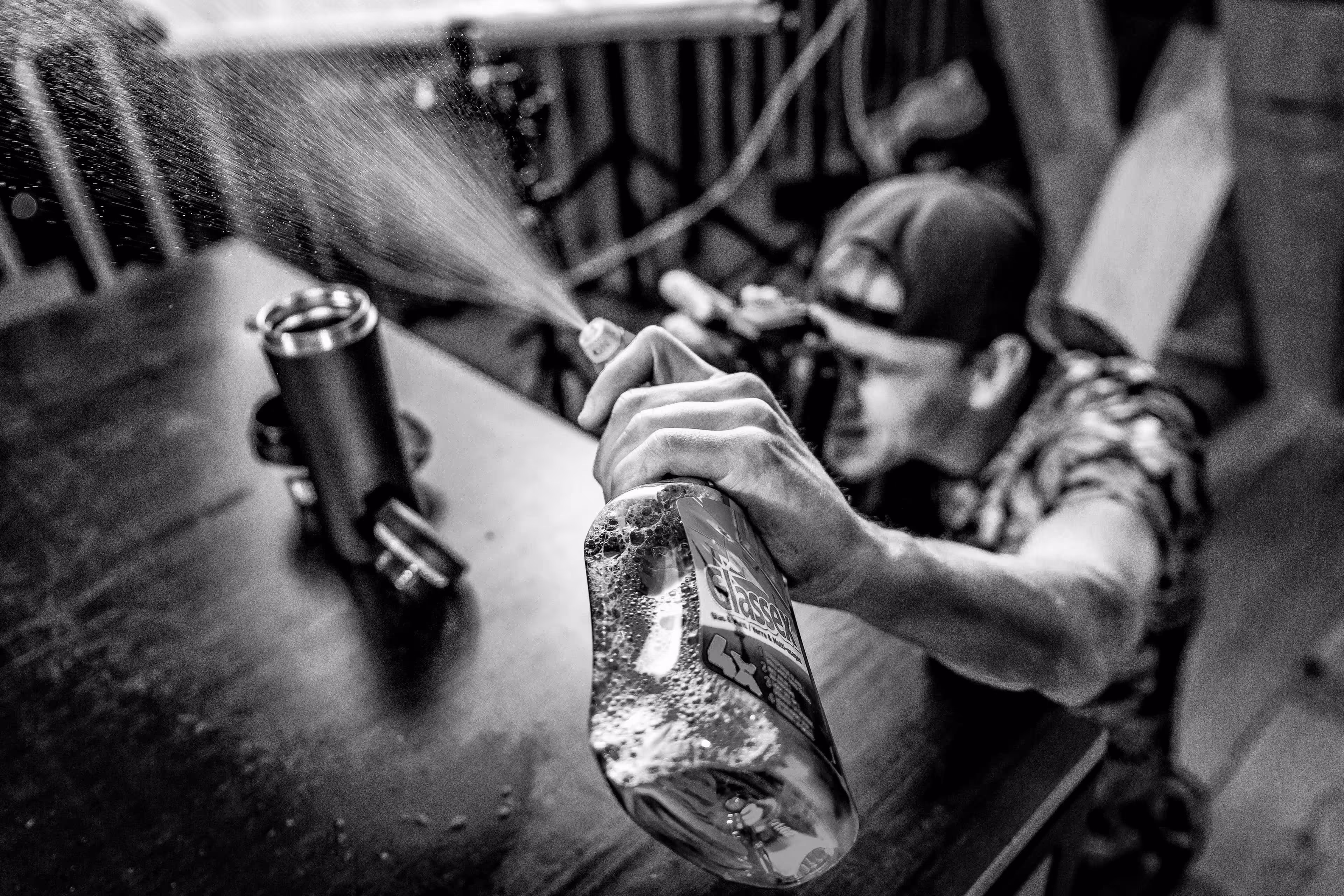 Person spraying glass cleaner onto a stainless steel travel mug on a wooden table.