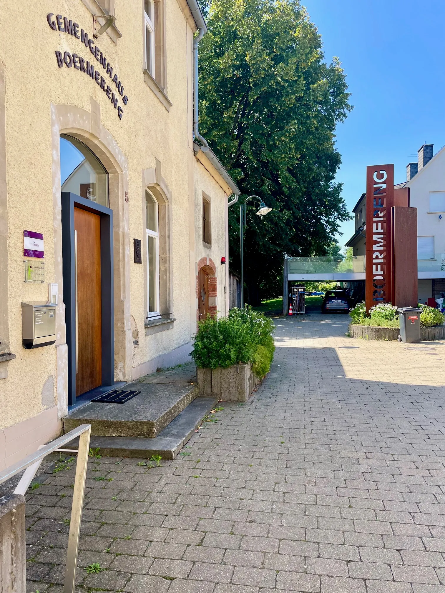 Sidewalk leading past a beige building with a wooden door and sign 'Gemeinckhaus Boermereng,' with a metal vertical sign and parked car in the background under a blue sky.