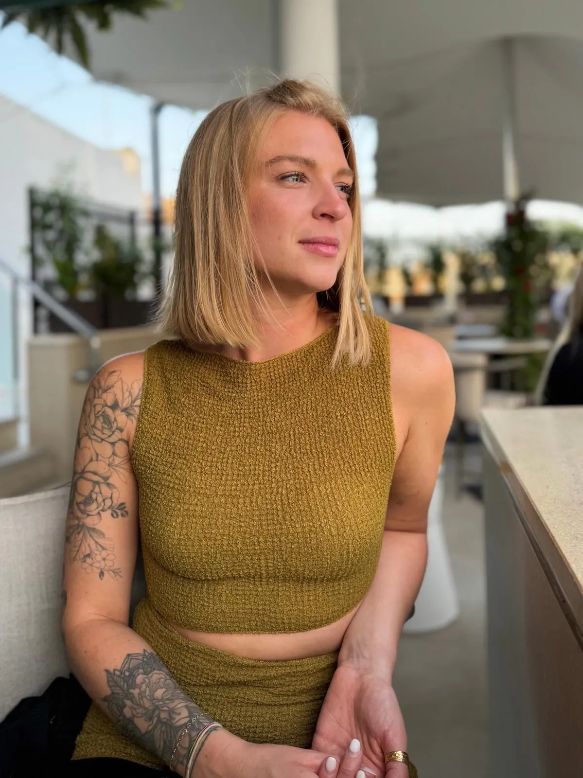 Blonde woman with floral tattoos on her left arm wearing a textured olive green sleeveless crop top, sitting outdoors at a cafe.