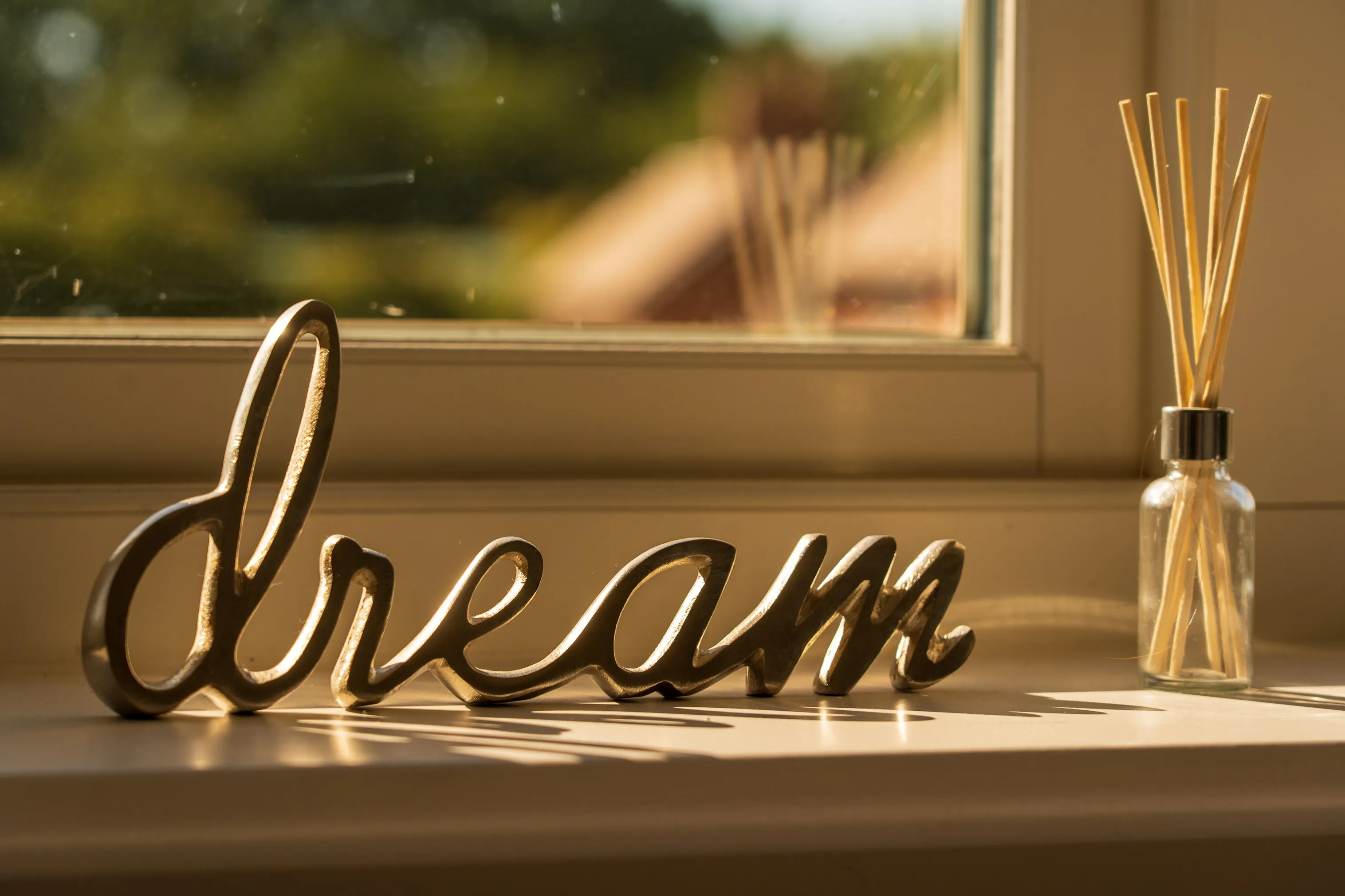 Metal decorative word 'dream' on a windowsill with a glass diffuser bottle beside it, bathed in warm sunlight.