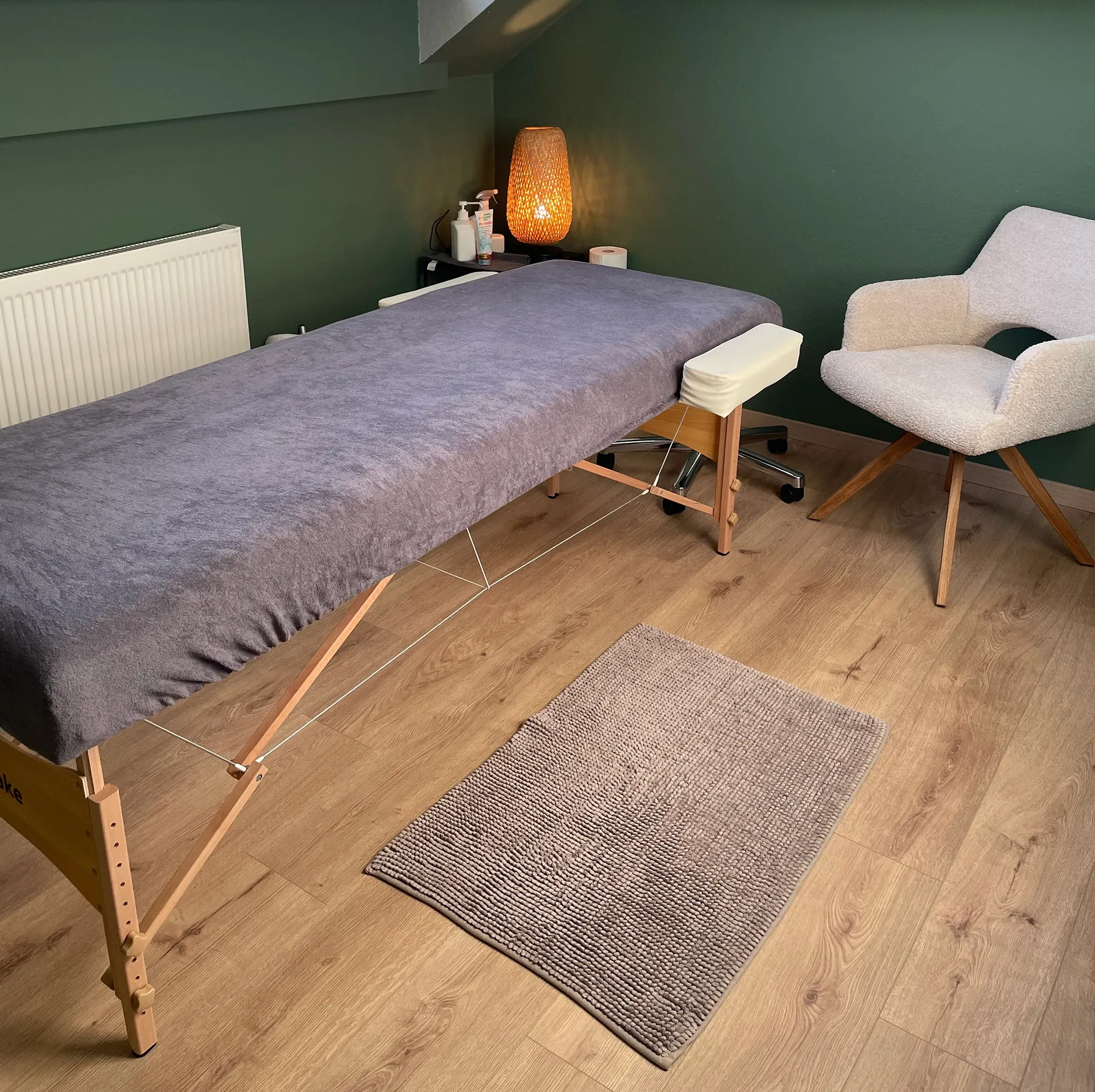 Massage therapy room with a covered massage table, cushioned chair, floor rug, and dim lamp on a small shelf against a green wall.