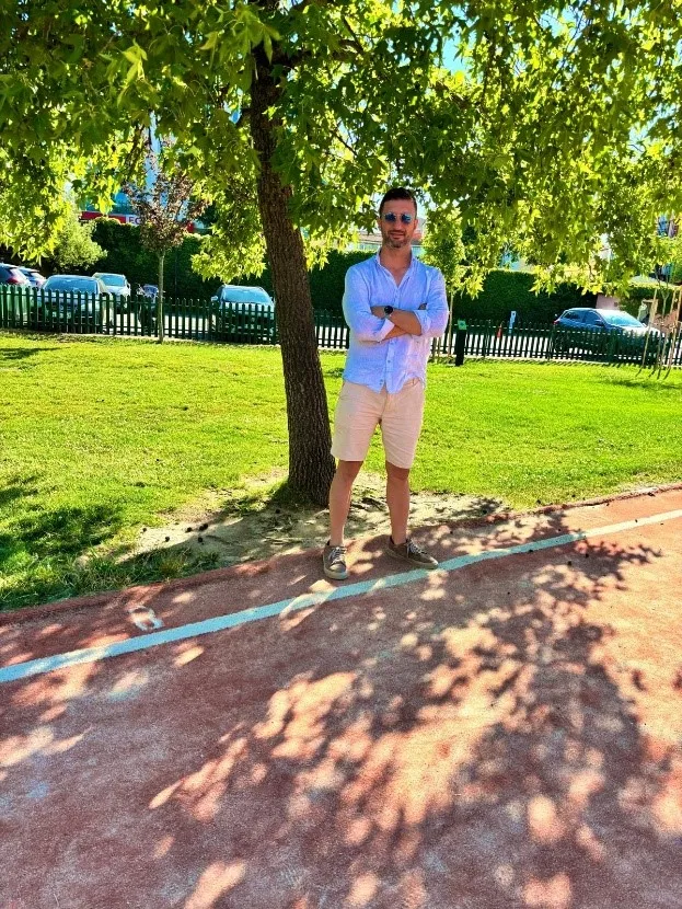 Güneş gözlüğü takan, açık mavi gömlek ve bej şort giyen bir adam, ağaçların ve çitin önünde kollarını kavuşturmuş halde dışarıda duruyor.