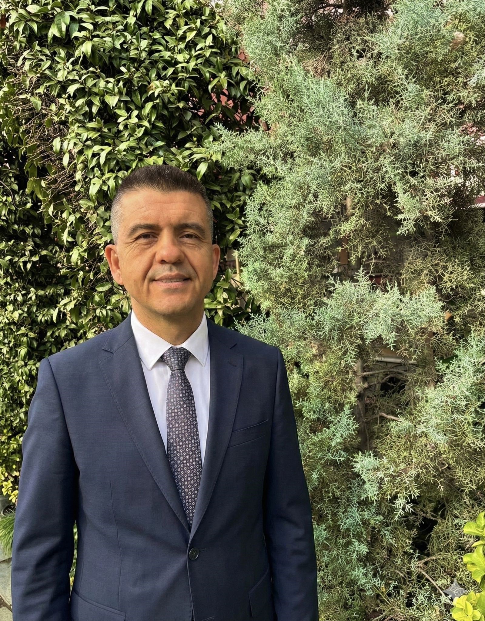 Koyu mavi takım elbise ve desenli kravat giymiş bir adam, arka planda yeşil yapraklı çalılıklar bulunan açık havada duruyor.