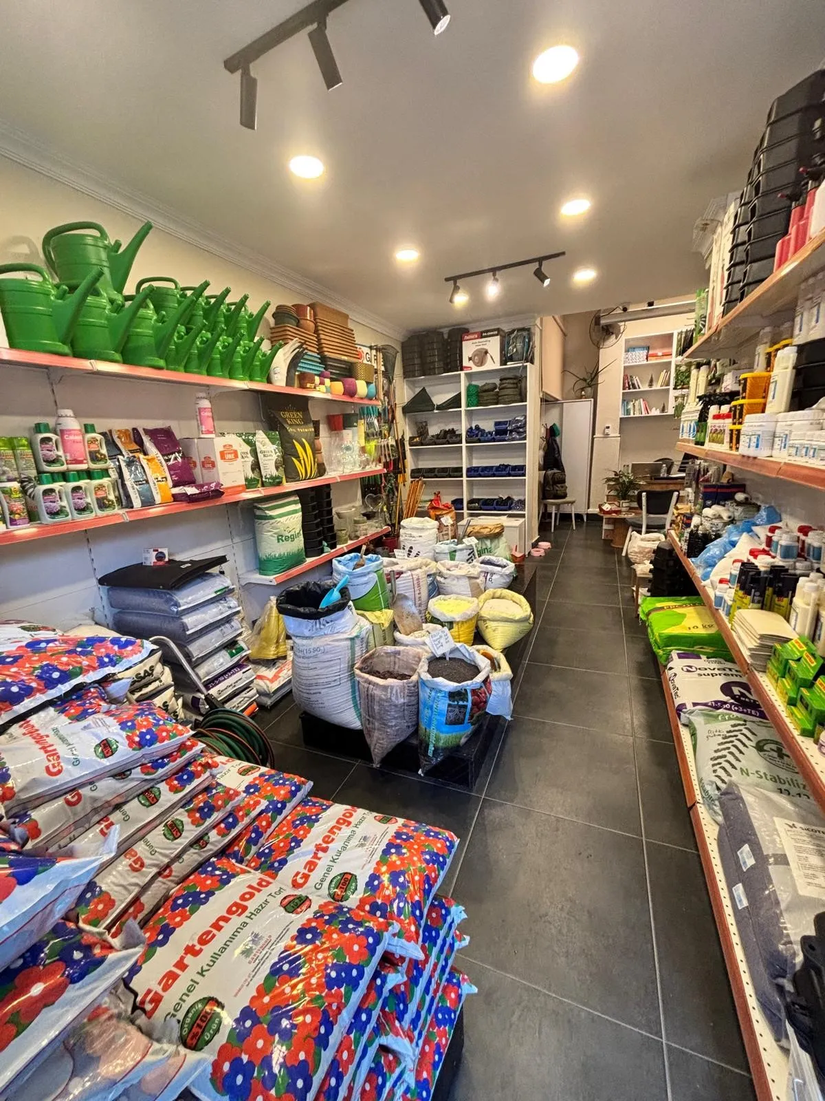 Raflarında sulama kapları, gübreler, toprak torbaları, bahçe aletleri ve çeşitli bahçe ürünleri bulunan bir bahçe malzemeleri mağazasının iç görünümü.