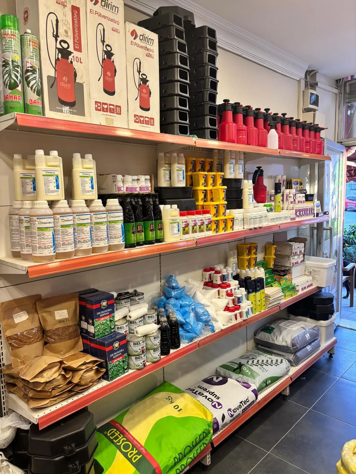 Mağazada sprey şişeleri, gübre torbaları, kimyasal şişeler ve tohum paketleri de dahil olmak üzere tarım ürünleriyle dolu raflar.
