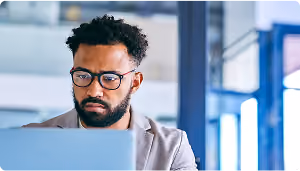 Man with glasses and curly hair working on a laptop in an office with large windows.