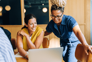 Two women staring at a laptop computer smiling while one is seated and the other is standing with hands resting on chairs.