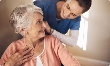 Smiling elderly woman in a pink cardigan being gently cared for by a smiling female healthcare worker in blue scrubs.