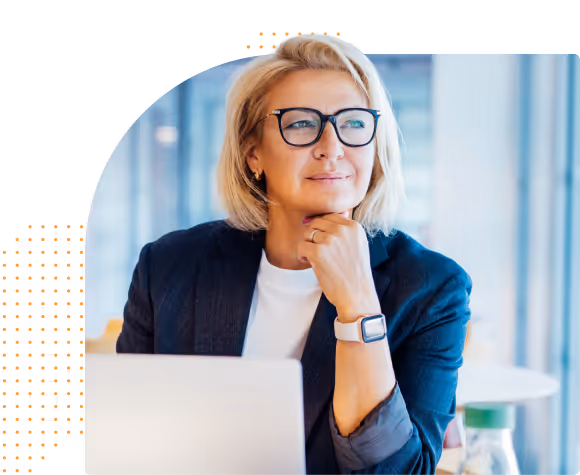 Middle-aged woman with glasses sitting thoughtfully at a desk with a laptop in a modern office.