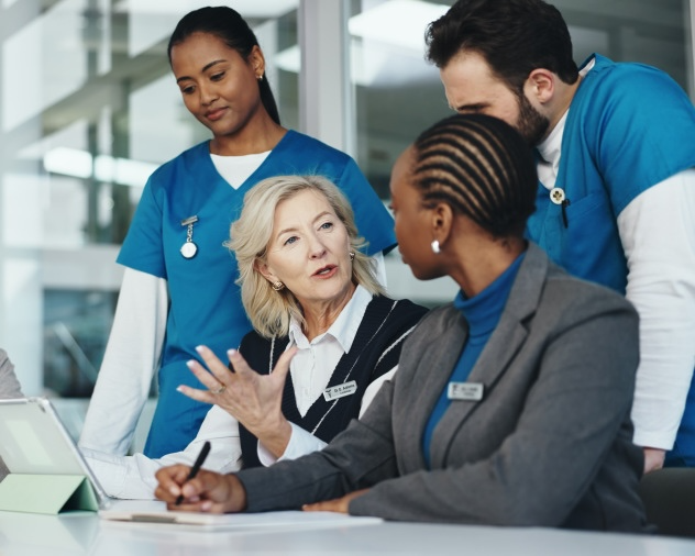 Four healthcare professionals, two in blue scrubs and two in business attire, engaged in a discussion around a table.
