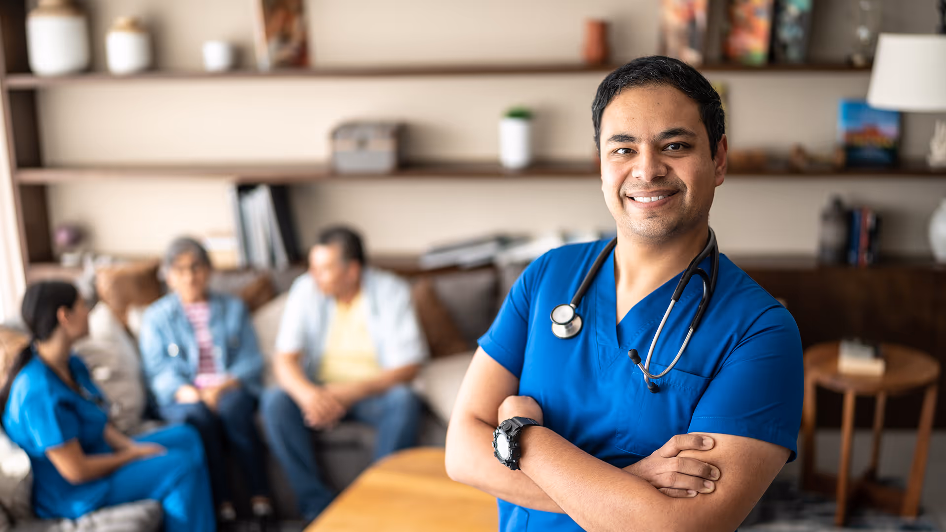 Smiling male nurse in blue scrubs with stethoscope standing with arms crossed in a living room with elderly couple and female caregiver in background.