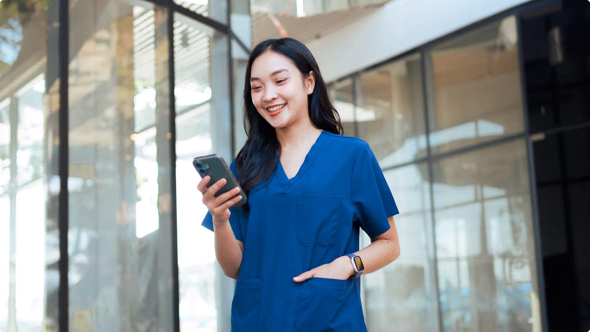 Smiling nurse wearing blue scrubs looking at her smartphone in a bright modern hallway.