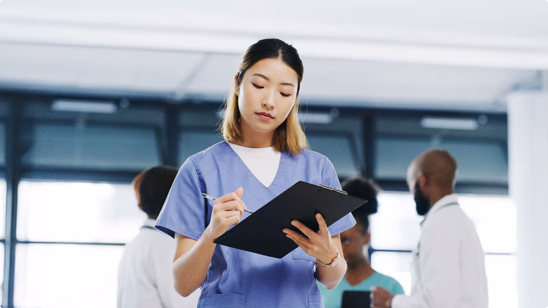 Healthcare professional in blue scrubs writing on a clipboard with medical staff in the background.