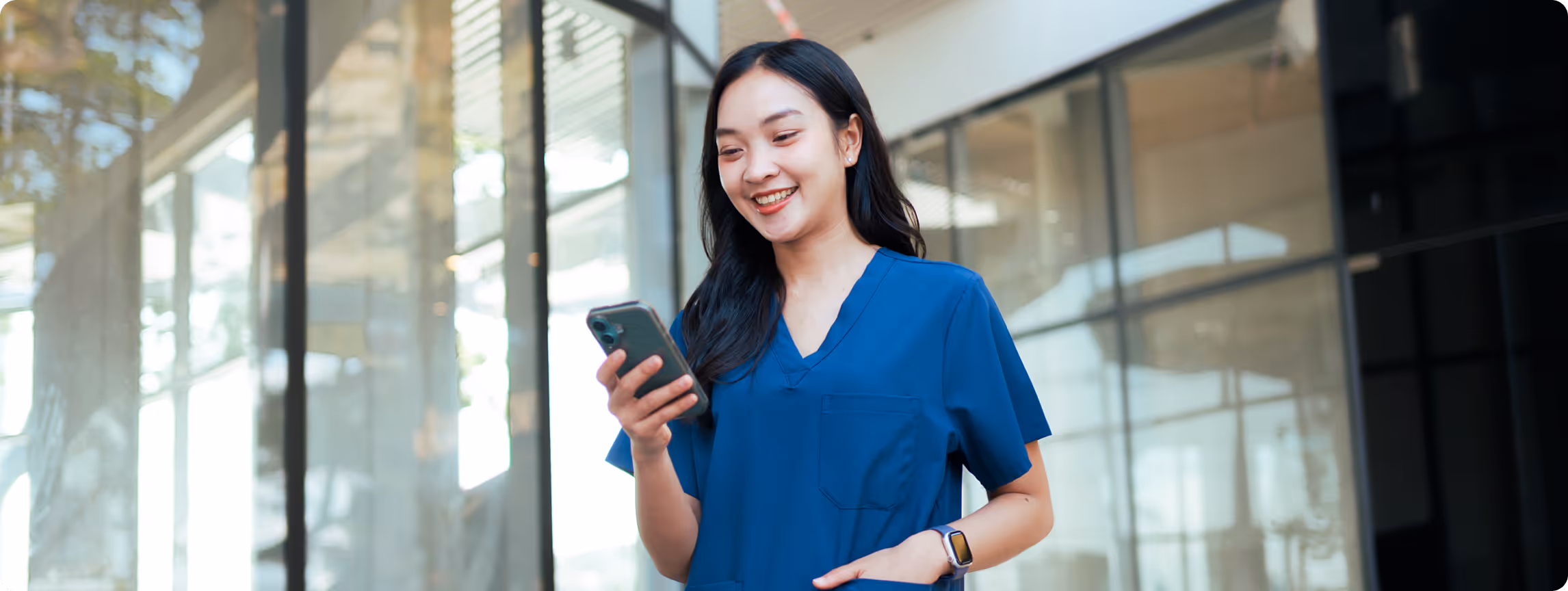 Smiling woman in blue scrubs looking at her smartphone in a modern glass-walled building.