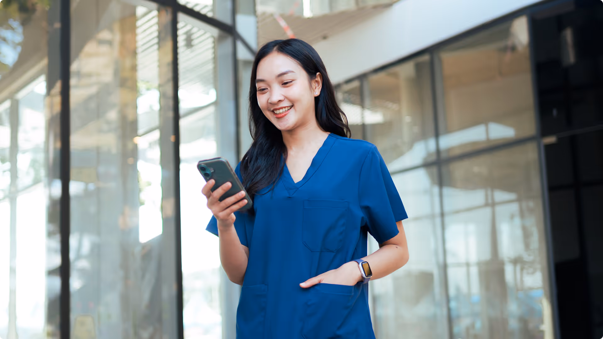 Smiling nurse in blue scrubs looking at a smartphone while standing indoors.
