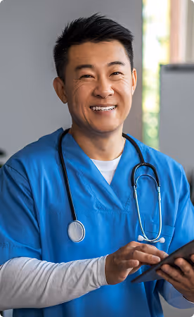 Smiling male healthcare professional wearing blue scrubs and a stethoscope, using a tablet device indoors.