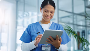 Smiling nurse in blue scrubs using a tablet in a bright medical facility.