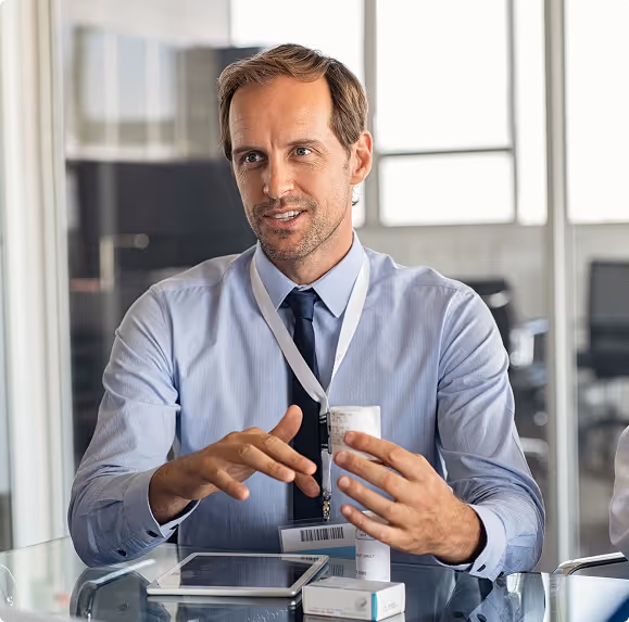 Man in a blue shirt and tie holding and explaining a medicine bottle in an office setting with a tablet and medicine box on the glass table.