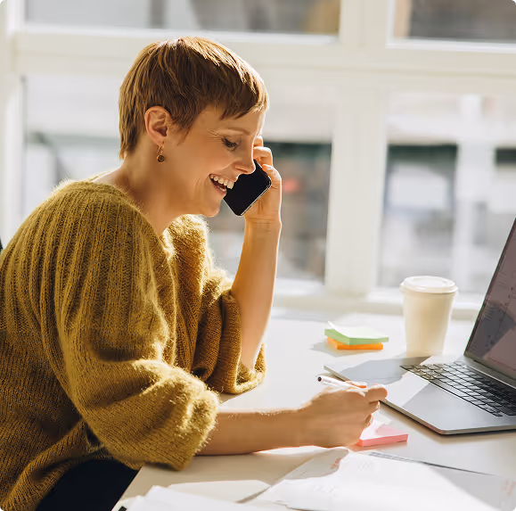 Woman with short hair wearing a mustard sweater smiling while talking on a smartphone and writing notes at a desk with a laptop and coffee cup.