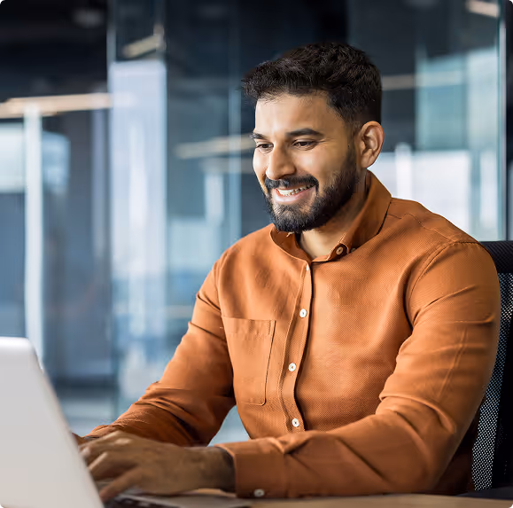 Smiling man in a brown shirt typing on a laptop in a modern office.