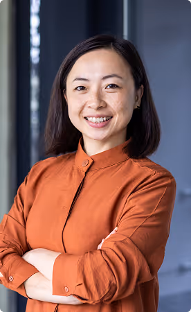 Smiling woman with shoulder-length dark hair wearing an orange buttoned shirt, standing with arms crossed indoors.