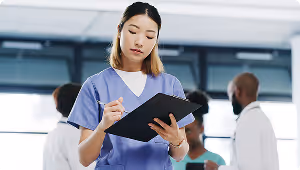 Healthcare professional in scrubs writing on a clipboard with blurred colleagues in the background.