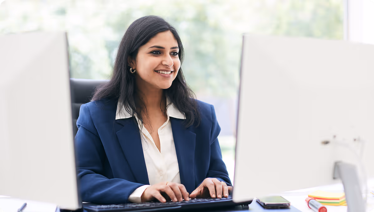 Smiling woman in business attire working on a computer at a desk with office supplies nearby.