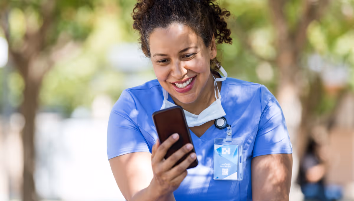 Smiling female healthcare worker in blue scrubs looking at a smartphone outdoors.