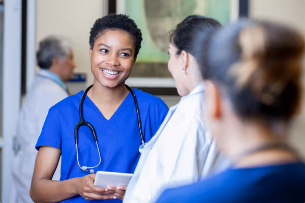 Smiling female nurse in blue scrubs holding a tablet and talking with a female doctor in a medical setting.