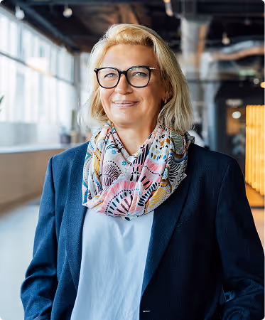 Smiling middle-aged woman with blonde hair and glasses wearing a colorful patterned scarf and dark blazer in a modern office setting.