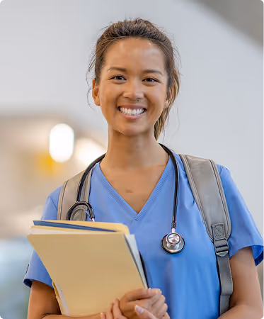 Smiling student nurse in blue scrubs holding folders with a stethoscope around her neck and backpack on shoulders.