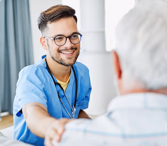 Smiling male healthcare professional in blue scrubs with stethoscope reassuring an elderly patient by placing a hand on their shoulder.