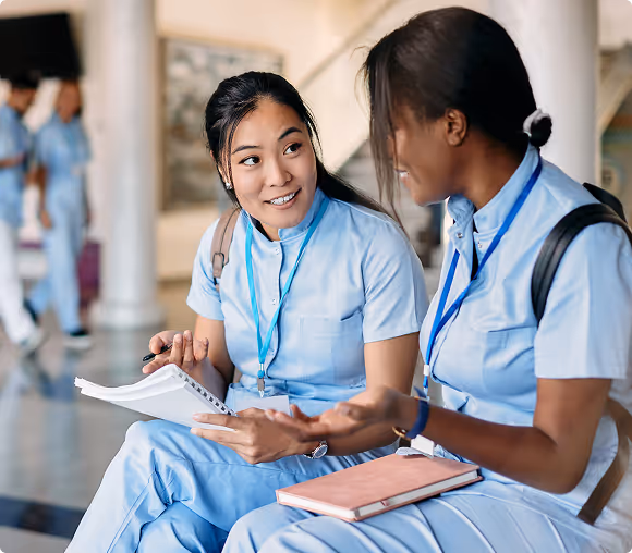 Two female medical professionals in light blue scrubs sitting and discussing notes in a hallway.