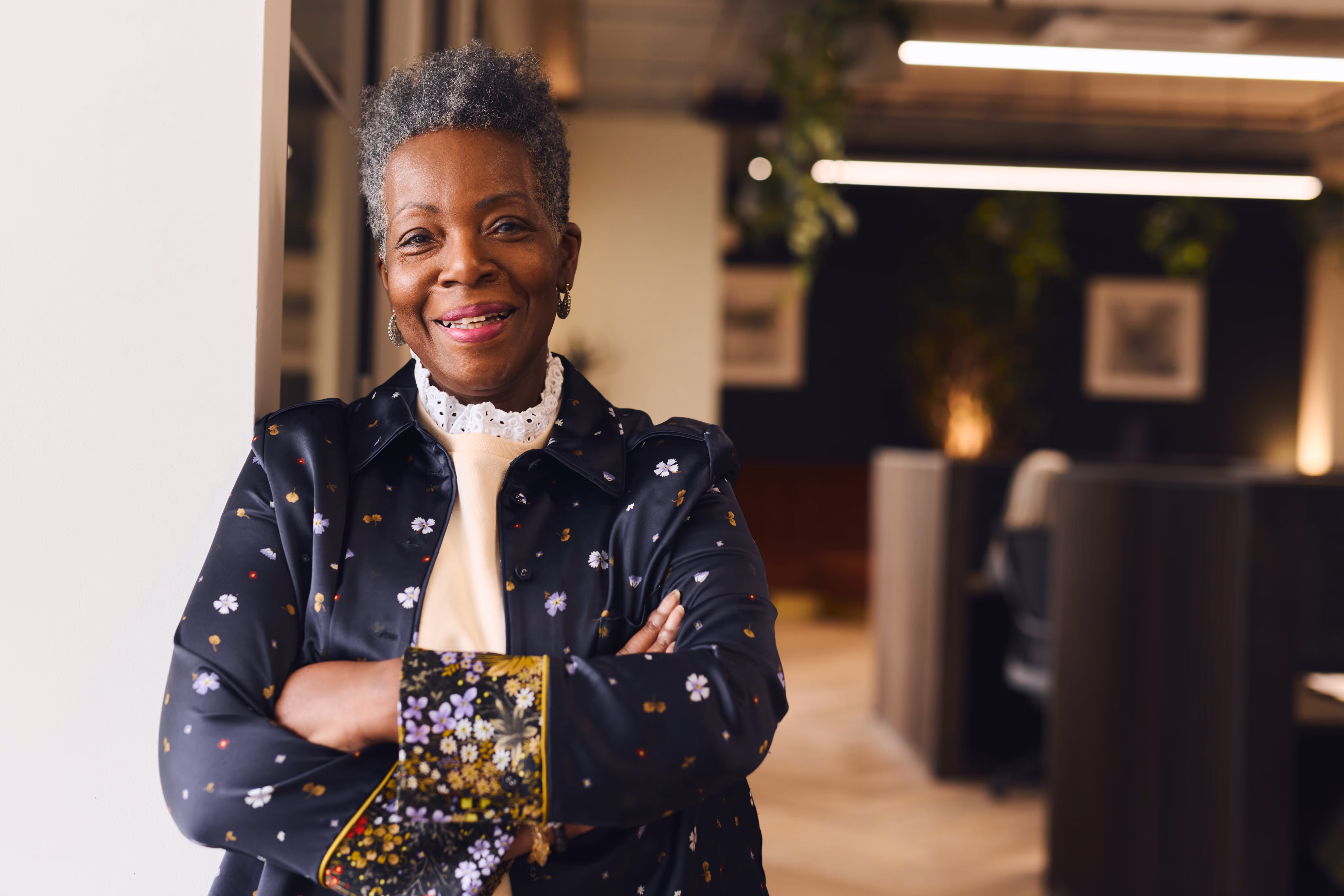 Smiling older woman with short gray hair wearing a floral black jacket, standing with crossed arms in a modern office.