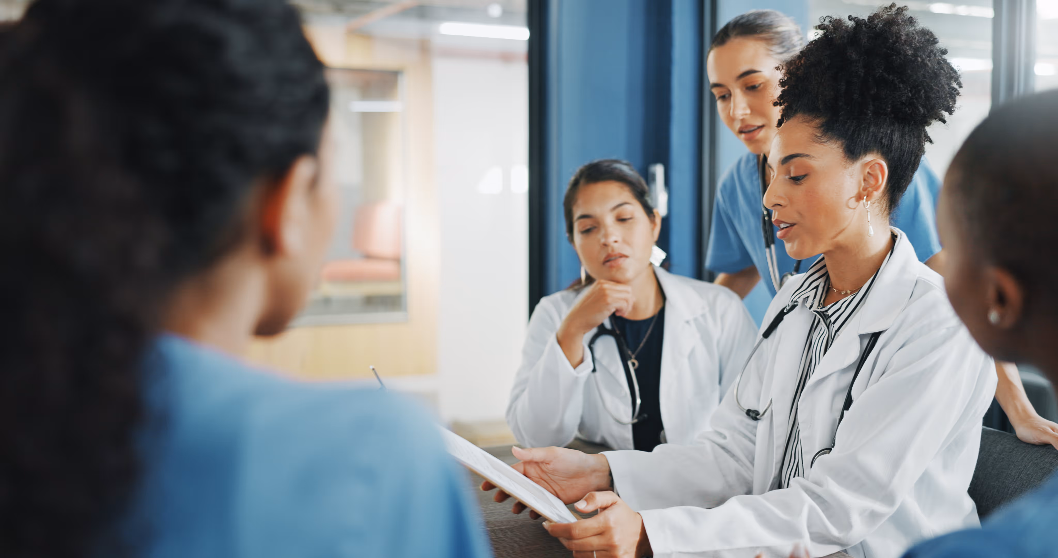 Diverse group of healthcare workers in white coats and scrubs discussing together around a table.