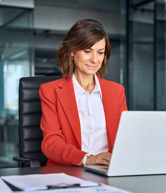 Professional woman in a red blazer working on a laptop at an office desk.