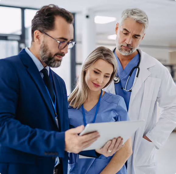 Three healthcare professionals, two in scrubs and one in a suit, examining a tablet together in a modern hospital hallway.