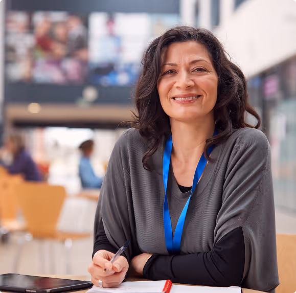 Smiling woman wearing a blue lanyard seated at a desk with a notebook and pen, in a bright indoor setting.