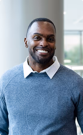 Smiling man with short hair and beard wearing a blue sweater over a white collared shirt indoors.