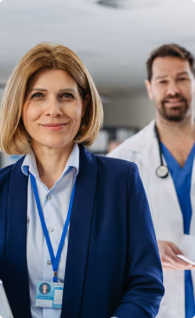 Smiling female healthcare professional in a navy blazer with an ID badge, with a male doctor in scrubs and a white coat in the background.