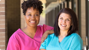 Two smiling female healthcare workers wearing pink and blue scrubs standing side by side in a hallway.