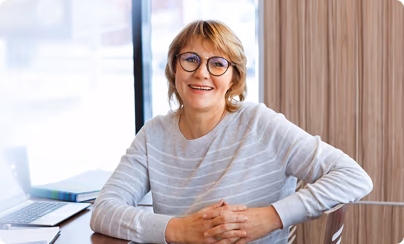 Smiling middle-aged woman with short blonde hair and glasses sitting at a table with a laptop and notebooks in a bright room.