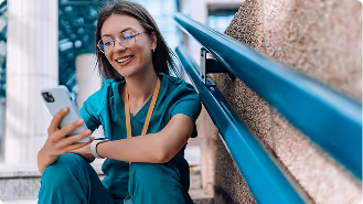 Smiling female healthcare worker in green scrubs sitting on outdoor steps looking at smartphone.