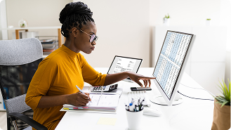 Woman in glasses and yellow sweater working with documents and pointing at data on a computer screen at a desk.